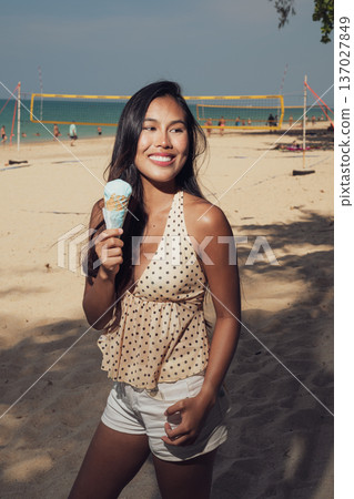 Smiling woman holding ice cream cone on sandy beach with volleyball court behind Smiling woman holding ice cream cone on sandy beach with volleyball court behind 137027849