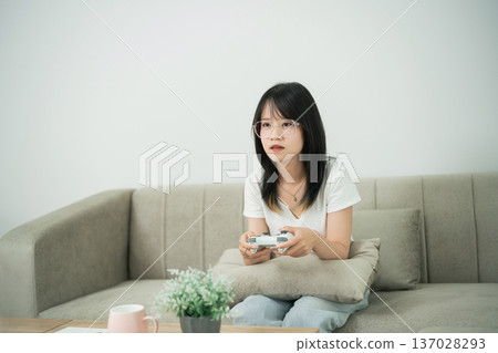 Young woman sitting on a sofa playing a video game with a controller in her hand, focused and enjoying leisure time indoors in a modern living space 137028293