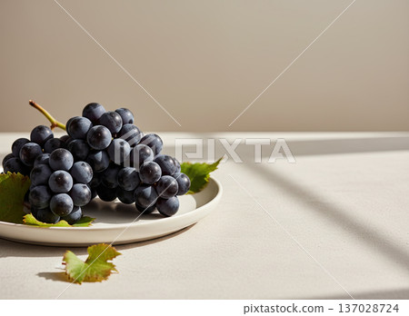 A bunch of dark purple grapes with green leaves resting on a white plate, beautifully lit by natural sunlight. 137028724