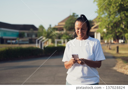 Smiling Mature Scandinavian Tourist Woman Walking Outdoors on Summer Holiday Texting With Phone Smiling Mature Scandinavian Tourist Woman Walking Outdoors on Summer Holiday Texting With Phone 137028824