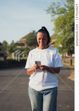 Smiling Mature Scandinavian Tourist Woman Walking Outdoors on Summer Holiday Texting With Phone Smiling Mature Scandinavian Tourist Woman Walking Outdoors on Summer Holiday Texting With Phone 137028825