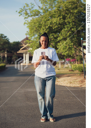 Smiling Mature Scandinavian Tourist Woman Walking Outdoors on Summer Holiday Texting With Phone Smiling Mature Scandinavian Tourist Woman Walking Outdoors on Summer Holiday Texting With Phone 137028827