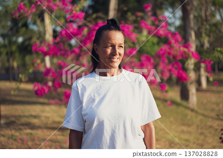 Smiling Mature Woman Standing in Tropical Garden with Pink Flowers Smiling Mature Woman Standing in Tropical Garden with Pink Flowers 137028828
