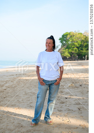 Mature woman standing on tropical beach wearing 90s style normcore fashion jeans and white t-shirt Mature woman standing on tropical beach wearing 90s style normcore fashion jeans and white t-shirt 137028856