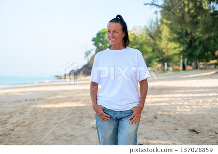 Mature woman standing on tropical beach wearing 90s style normcore fashion jeans and white t-shirt Mature woman standing on tropical beach wearing 90s style normcore fashion jeans and white t-shirt 137028859