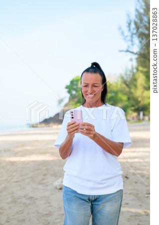 Mature woman standing on tropical beach wearing 90s style normcore fashion jeans and white t-shirt Mature woman standing on tropical beach wearing 90s style normcore fashion jeans and white t-shirt 137028863