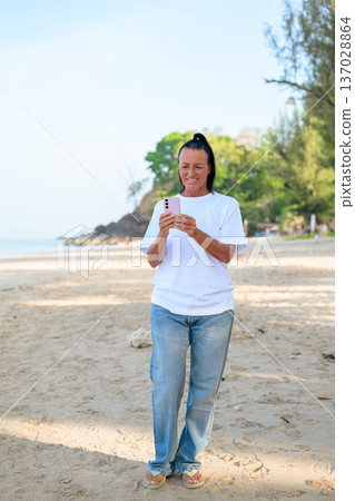 Mature woman standing on tropical beach wearing 90s style normcore fashion jeans and white t-shirt Mature woman standing on tropical beach wearing 90s style normcore fashion jeans and white t-shirt 137028864