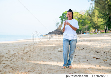 Mature woman standing on tropical beach wearing 90s style normcore fashion jeans and white t-shirt Mature woman standing on tropical beach wearing 90s style normcore fashion jeans and white t-shirt 137028865