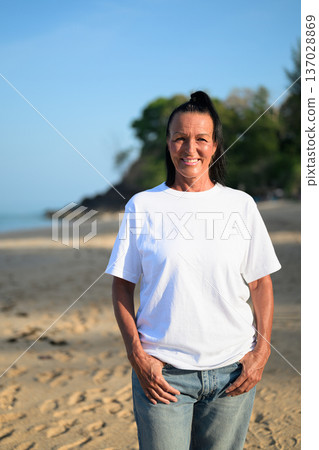 Mature woman standing on tropical beach wearing 90s style normcore fashion jeans and white t-shirt Mature woman standing on tropical beach wearing 90s style normcore fashion jeans and white t-shirt 137028869
