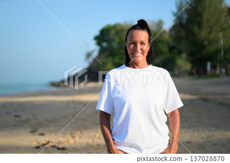 Mature woman standing on tropical beach wearing 90s style normcore fashion jeans and white t-shirt Mature woman standing on tropical beach wearing 90s style normcore fashion jeans and white t-shirt 137028870