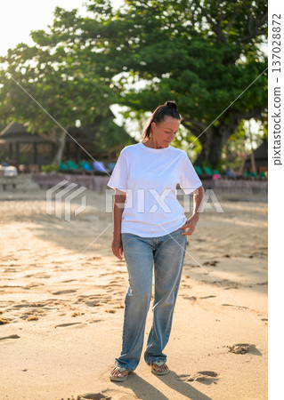 Mature woman standing on tropical beach wearing 90s style normcore fashion jeans and white t-shirt Mature woman standing on tropical beach wearing 90s style normcore fashion jeans and white t-shirt 137028872