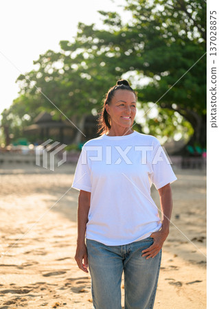 Mature woman standing on tropical beach wearing 90s style normcore fashion jeans and white t-shirt Mature woman standing on tropical beach wearing 90s style normcore fashion jeans and white t-shirt 137028875