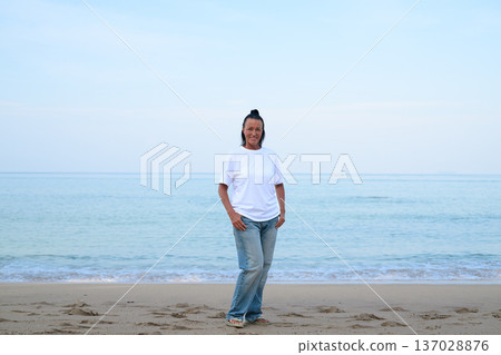 Mature woman standing on tropical beach wearing 90s style normcore fashion jeans and white t-shirt 137028876