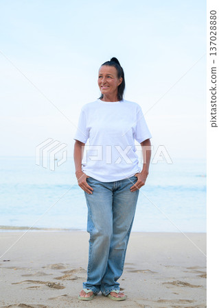 Mature woman standing on tropical beach wearing 90s style normcore fashion jeans and white t-shirt Mature woman standing on tropical beach wearing 90s style normcore fashion jeans and white t-shirt 137028880