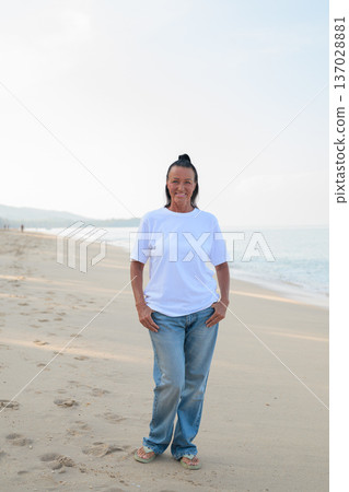 Mature woman standing on tropical beach wearing 90s style normcore fashion jeans and white t-shirt 137028881