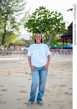Mature woman standing on tropical beach wearing 90s style normcore fashion jeans and white t-shirt 137028887