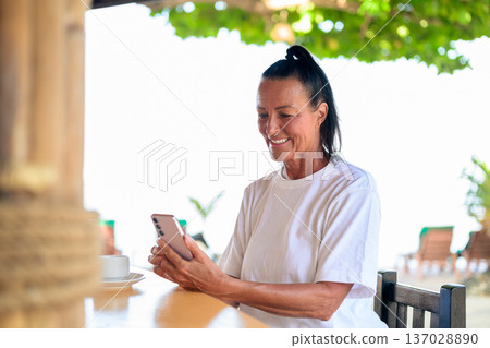 Senior woman texting on phone while enjoying coffee in tropical setting 137028890