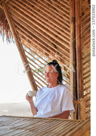 Smiling Mature Woman Drinking Coffee on Beachside Bamboo Terrace Smiling Mature Woman Drinking Coffee on Beachside Bamboo Terrace 137028902