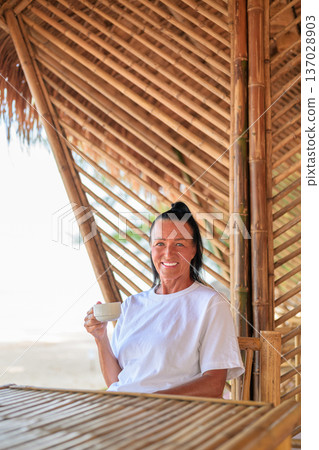 Smiling Mature Woman Drinking Coffee on Beachside Bamboo Terrace Smiling Mature Woman Drinking Coffee on Beachside Bamboo Terrace 137028903