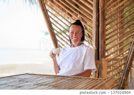 Smiling Mature Woman Drinking Coffee on Beachside Bamboo Terrace Smiling Mature Woman Drinking Coffee on Beachside Bamboo Terrace 137028904