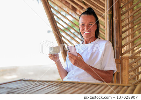 Smiling Senior Woman Using Smartphone and Drinking Coffee on Tropical Terrace Smiling Senior Woman Using Smartphone and Drinking Coffee on Tropical Terrace 137028907