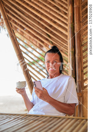 Smiling Senior Woman Using Smartphone and Drinking Coffee on Tropical Terrace 137028908