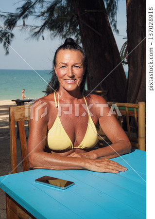 Confident senior woman in bikini relaxing at seaside cafe table with smartphone Confident senior woman in bikini relaxing at seaside cafe table with smartphone 137028918