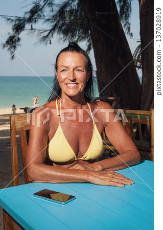Confident senior woman in bikini relaxing at seaside cafe table with smartphone Confident senior woman in bikini relaxing at seaside cafe table with smartphone 137028919