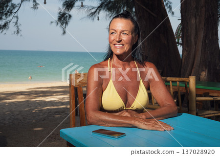 Confident senior woman in bikini relaxing at seaside cafe table with smartphone Confident senior woman in bikini relaxing at seaside cafe table with smartphone 137028920