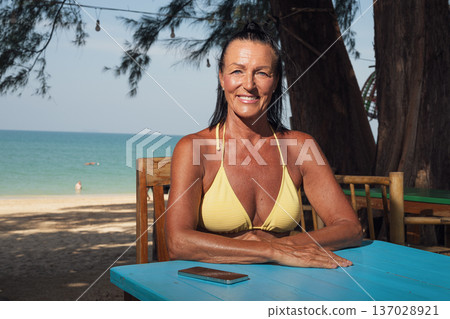 Confident senior woman in bikini relaxing at seaside cafe table with smartphone Confident senior woman in bikini relaxing at seaside cafe table with smartphone 137028921