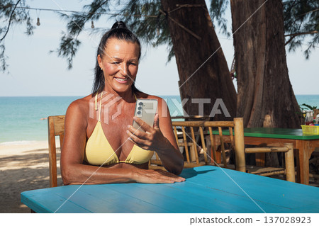 Smiling Fit Senior Woman Using Smartphone at Beach Cafe 137028923