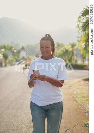 Smiling Mature Scandinavian Tourist Woman Walking Outdoors on Summer Holiday Texting With Phone 137028986