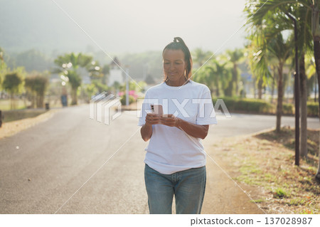 Smiling Mature Scandinavian Tourist Woman Walking Outdoors on Summer Holiday Texting With Phone Smiling Mature Scandinavian Tourist Woman Walking Outdoors on Summer Holiday Texting With Phone 137028987