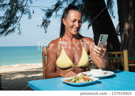 Active senior woman checks her phone while enjoying seafood dish by the ocean in restaurant cafe 137029005