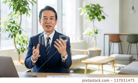 一位中年日本商人正在明亮的辦公室裡嚴肅地講話,並伴隨著手勢。 一位中年日本商人正在明亮的辦公室裡嚴肅地講話,並伴隨著手勢。 137031475