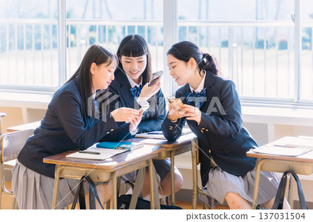 A high school girl talking to her classmate while looking at her smartphone in the classroom 137031504