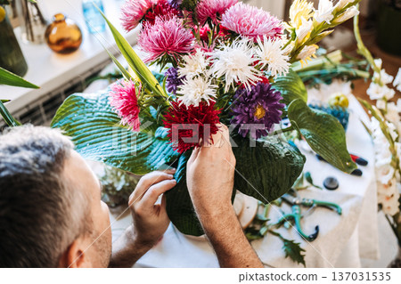 man arrange aster flowers and large green leaves in vintage vase. Stress management for men, therapeutic floristry, cortisol reduction, mindful craftsmanship, male wellness. 137031535