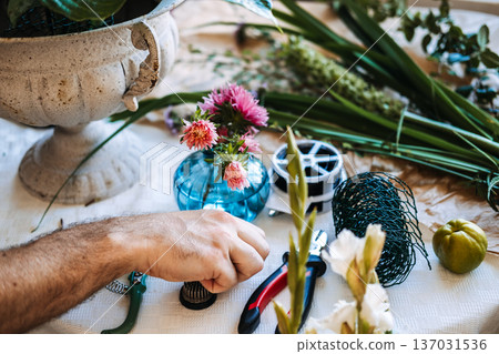 Man places small flower frog on table next to floral tools and aster blooms. Artisanal floral design, master florist at work, handcrafted aesthetic, boutique floral studio. Man places small flower frog on table next to floral tools and aster blooms. Artisanal floral design, master florist at work, handcrafted aesthetic, boutique floral studio. 137031536
