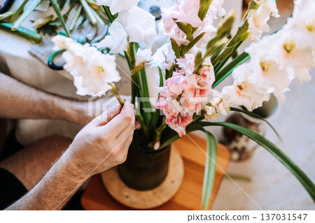 Man arranges peach gladiolus stems into dark glass vase on white table. Indoor flower styling, botanical living spaces, urban jungle lifestyle. 137031547