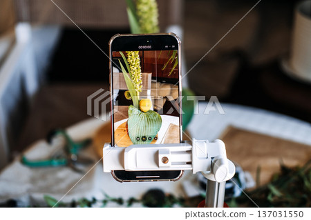Smartphone mounted on tripod records video of floral arrangement in glass bottle. floral workshop, teaching craft, hobbyist community, skill-sharing, micro-influencer, educational social media Smartphone mounted on tripod records video of floral arrangement in glass bottle. floral workshop, teaching craft, hobbyist community, skill-sharing, micro-influencer, educational social media 137031550