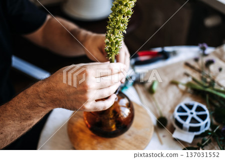 Hands place tall green flower stalk into glass vase on table. Japanese floral arrangement, minimalist aesthetics, meditative art, zen lifestyle, precision floristry, cultural heritage. Hands place tall green flower stalk into glass vase on table. Japanese floral arrangement, minimalist aesthetics, meditative art, zen lifestyle, precision floristry, cultural heritage. 137031552