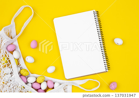 Colored Easter eggs in a mesh bag and a blank notepad on a yellow background. Colored Easter eggs in a mesh bag and a blank notepad on a yellow background. 137031947