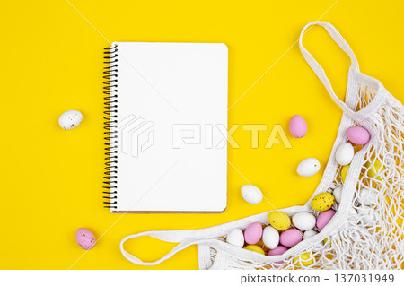 Colored Easter eggs in a mesh bag and a blank notepad on a yellow background. 137031949