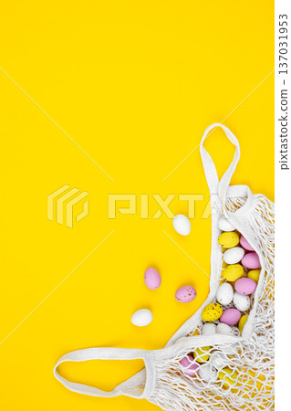 Colored Easter eggs in a mesh bag on a yellow background, flat lay. 137031953