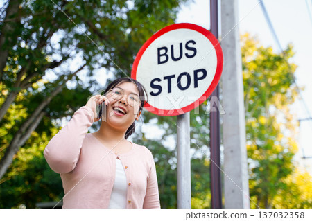 Young Asian woman talking on a smartphone while waiting at a bus stop. Young Asian woman talking on a smartphone while waiting at a bus stop. 137032358