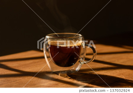 Warm cup of coffee steaming on a wooden table with dramatic sunlight filtering through window shadows Warm cup of coffee steaming on a wooden table with dramatic sunlight filtering through window shadows 137032924