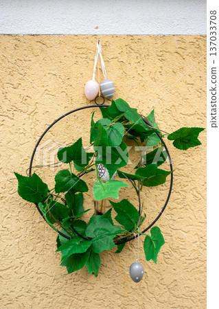 Simple Easter wreath made of green leaves and hanging pastel eggs on textured yellow wall. Spring holiday decoration and festive home design. 137033708
