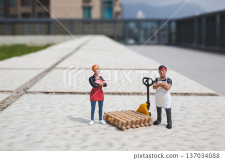 Miniature workers stand beside pallet and yellow hand truck Miniature workers stand beside pallet and yellow hand truck 137034088