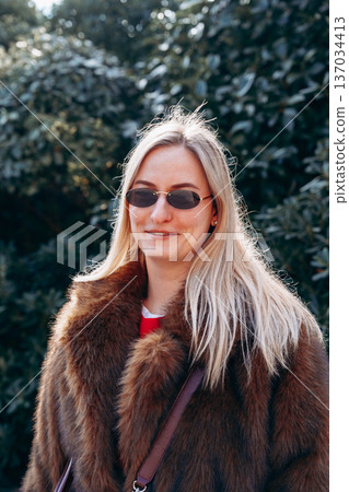 Woman wears sunglasses and a fur coat in a green garden during daytime 137034413