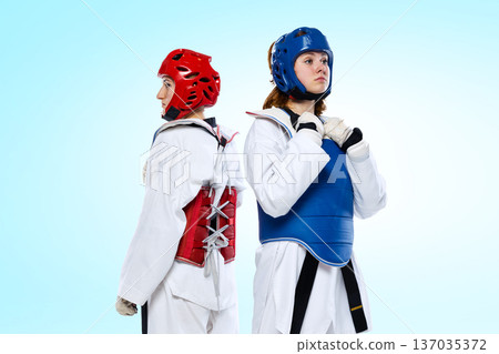 Female taekwondo athletes standing back to back in red and blue protective armor on light blue background. Female taekwondo athletes standing back to back in red and blue protective armor on light blue background. 137035372